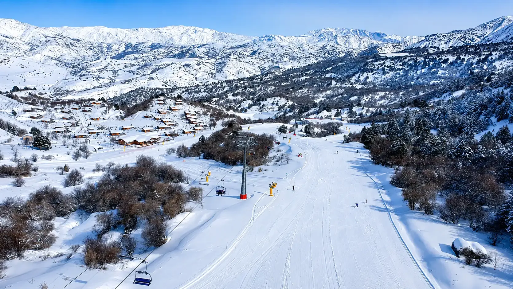 Amirsoy chalet village and chairlift beside a groomed winter piste under snow-covered ranges.