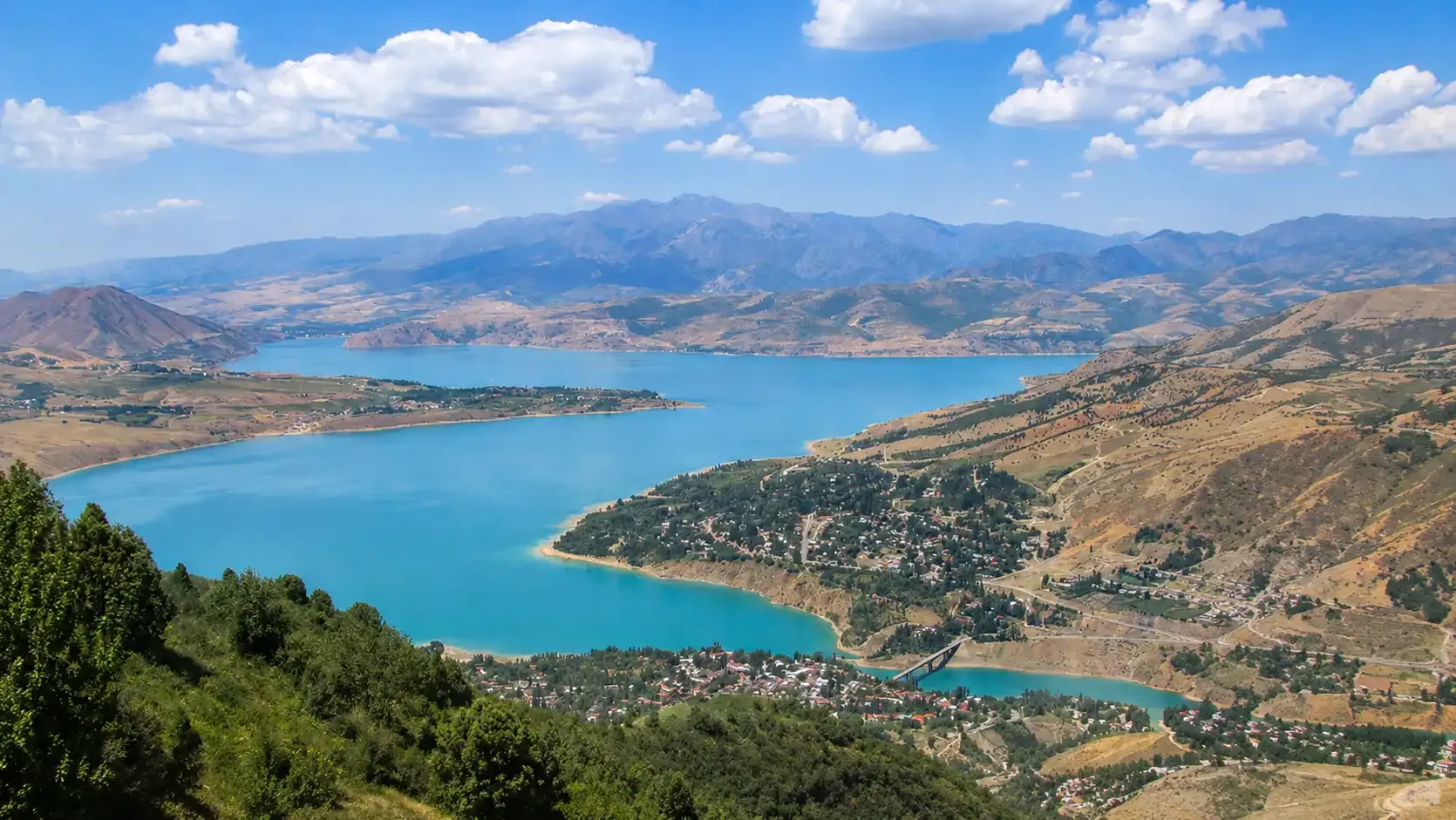 The turquoise Charvak reservoir seen from the ridge above, with a lakeside village and its bridge.