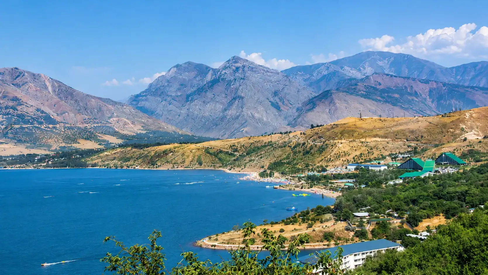 The Charvak shoreline in summer — beach, boats and resort roofs under bare rock ridges.